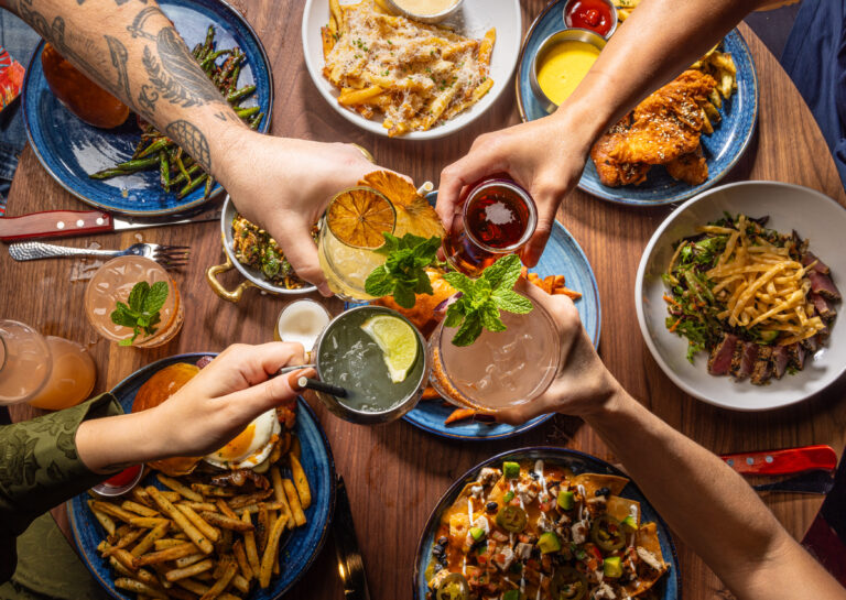 Top view of four people clinking their cocktails over a table full of American bar and grill food