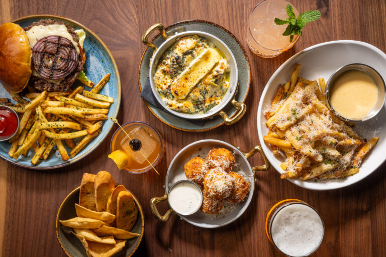 Arrangements of American bar and grill food, with plates of parmesan french fries, roasted vegetable gratin, and an open grilled onion burger